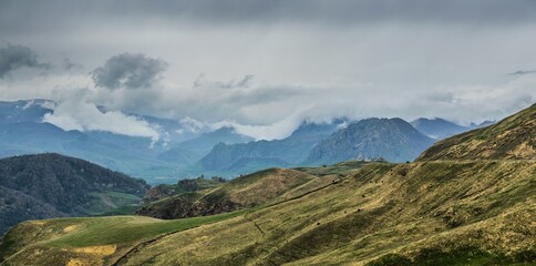 Panoramic landscape view over Gum-bashi mountain pass, with new green and old brown grass on cliffs, dark clouds before the storm, Сaucasus mountian range and Elbrus mountain. Karachay-Cherkessia