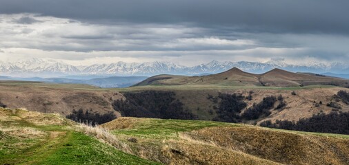 Panoramic landscape view over Gum-bashi mountain pass, with new green and old brown grass on cliffs, dark clouds before the storm, Сaucasus mountian range and Elbrus mountain. Karachay-Cherkessia