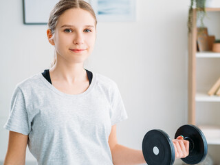Fototapeta na wymiar Child home sport. Fitness indoors. Wellbeing motivation. Athletic girl in sportswear training arm biceps muscles with dumbbell in light room interior.