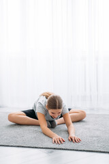 Fototapeta na wymiar Home sport. Kids fitness training. Morning workout. Active lifestyle. Athletic girl in sportswear doing stretching exercise on floor on light white window empty space background.