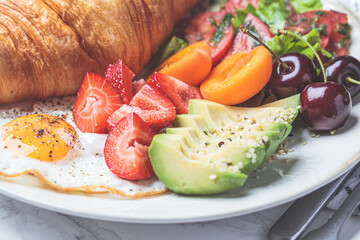 Breakfast plate with croissant, avocado, fried egg, salad and fruit.
