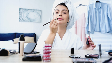 young woman in bathrobe with head wrapped in towel applying lipstick near table with cosmetics in bedroom