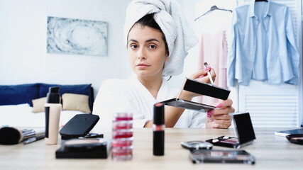 young woman in bathrobe sitting with cosmetic brush and eye shadows near table with decorative cosmetics in bedroom