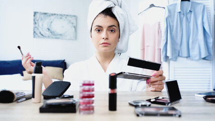 serious young woman in bathrobe sitting with cosmetic brush and eye shadows near table with decorative cosmetics in bedroom