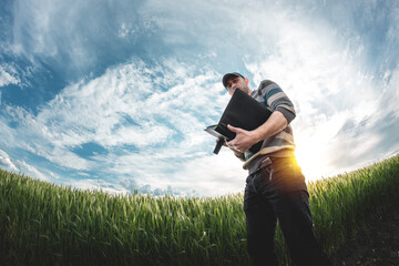 A young agronomist holds a folder in his hands on a green wheat field. A farmer makes notes on the background of agricultural land during sunset. Man in a cap with a folder of documents