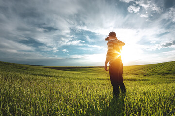 A young agronomist holds a folder in his hands on a green wheat field. A farmer makes notes on the background of agricultural land during sunset. Man in a cap with a folder of documents - obrazy, fototapety, plakaty