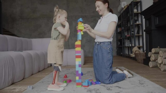 cheerful woman with amputee daughter near falling tower of building blocks