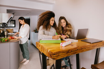 Long-haired Asian girl cutting a tomato in the kitchen while her friends are studying