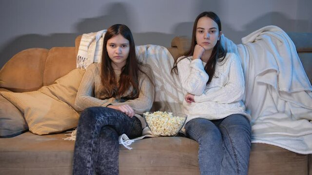Two teenage girls watching TV on sofa and eating popcorn at night