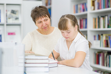 Fototapeta na wymiar Senior woman helps to girl with syndrome doing homework at library. Education for disabled children concept