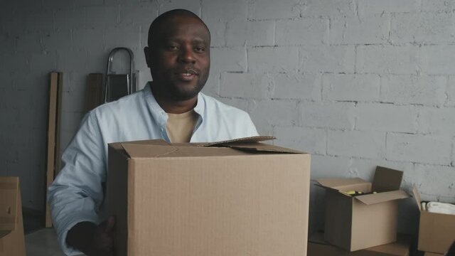 Slowmo medium shot of cheerful African-American man standing in his new house or apartment and receiving cardboard box from male mover or courier