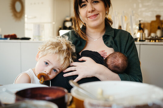 Candid moment of mother feeding newborn baby a the kitchen table and toddler resting his head - Powered by Adobe