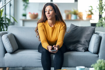 Worried exhausted woman looking her smartphone while waiting for news sitting on couch at home.