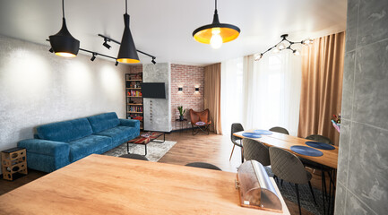Wide angle view of minimalist living room in beige, gray and blue tones with window, curtains, parquet, sofa, armchair, carpet, tables, rack, TV set. Pendant lamps in loft style in different versions.