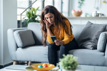 Worried exhausted woman looking her smartphone while waiting for news sitting on couch at home.