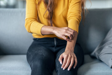 Young woman sitting on the couch while scratching her arm. Psoriasis concept.
