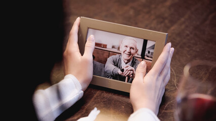 partial view of female hands holding photo frame with picture of elderly man at home