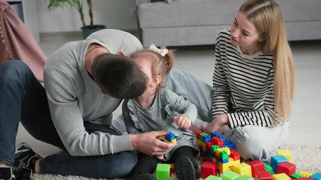 young loving family parents with daughter enjoy joint vacation kissing their female child and playing with colored plastic constructors in children room sitting on floor