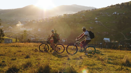 A married couple ride bicycles while traveling in the mountains, watching the sunset over the village