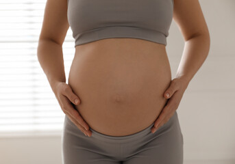 Pregnant young woman touching belly near window indoors, closeup