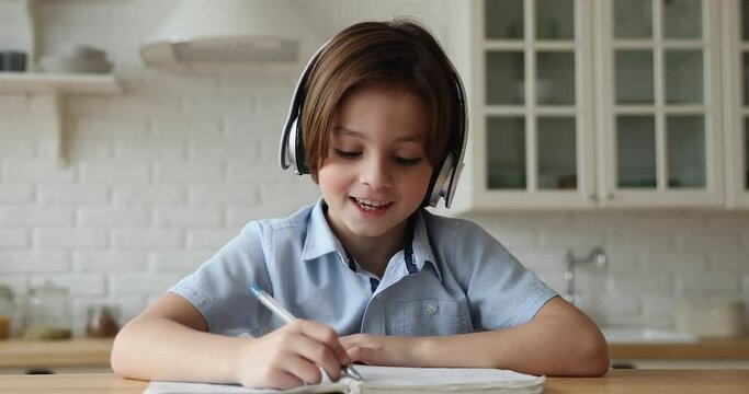 Pretty little 6s 7s boy wear headphones communicates to on-line tutor, writing notes in copybook studying subject use videocall app. Young gen and modern tech, internet, videoconference usage concept