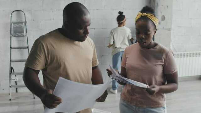 Medium shot of African-American man and woman looking at home remodeling plans and chatting while kids measuring wall in background