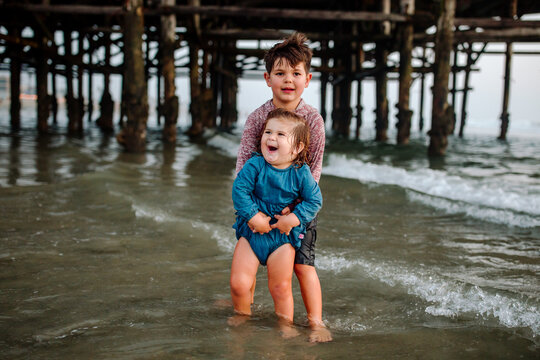 Brother steadies laughing sister in surf near pier 
