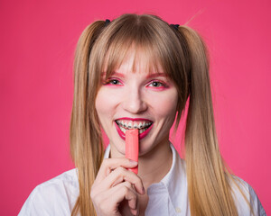 Portrait of a young woman with braces and bright makeup chewing gum on a pink background.