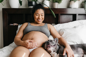 A portrait of a pregnant African American woman holding her french bulldog on the bed. 