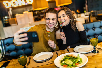Portrait of a joyful young couple taking a selfie while having lunch together at the cafe table indoors