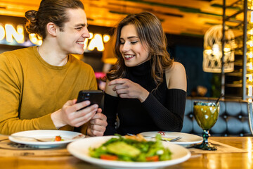 Cheerful young couple browsing internet on smartphone during romantic dinner at cozy cafe. Handsome boyfriend and girlfriend watching video together on cellphone while having lunch