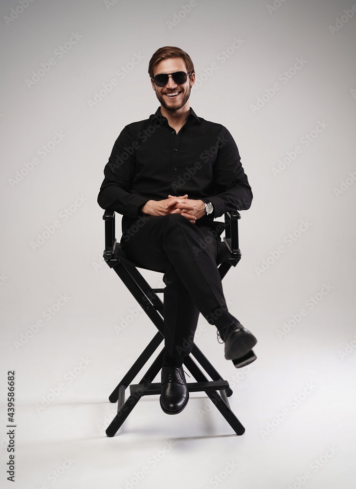 Wall mural portrait of a respectable man in a suit and sunglasses sitting on chair on a gray background.