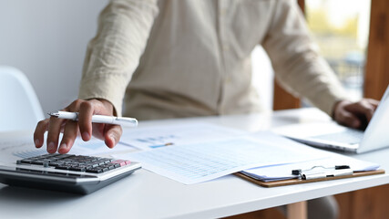 Hand with pen to use calculator and laptop keyboard, graphs and file folders on the desk.