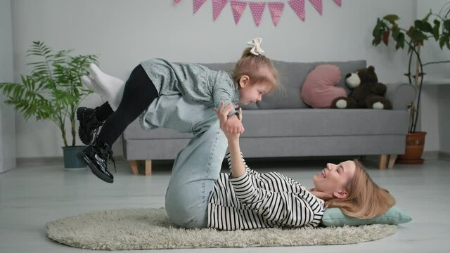 motherhood, happy female parent has fun with little adorable jock lying on her feet while playing airplane or doing physical exercises for mothers lying on floor at home