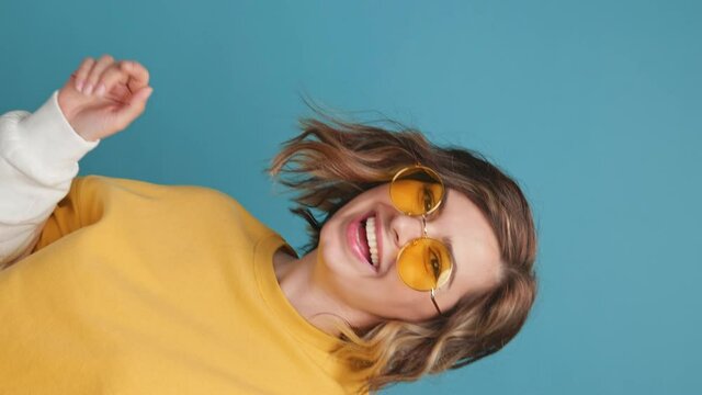 Pretty cute young woman appears unexpectedly in frame smiles affably in yellow sunglasses on blue background looks at camera vertical video for stories. People positive emotions concept
