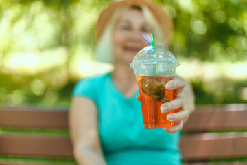 Summer beautiful happy woman hold glass of soda sitting on a wooden bench in city park, enjoying the rest. Healthy food concept