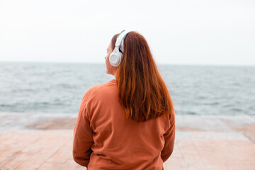 Back view of red hair woman wearing headphones listening music or podcast from smartphone...