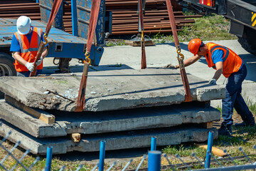 Two slingers unload concrete slabs on the street on a summer day. Workers in construction helmets...