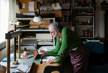 Grey-haired artisan woman using ipad in her small bookbinding workshop