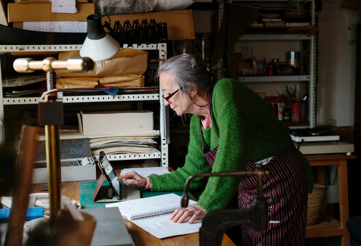 Senior woman in her artisan workshop checking device leaning on a desk - Powered by Adobe