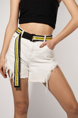 Cropped shot of slim girl in white ripped shorts, black crop top and striped canvas belt with D-rings buckle and lettering NICE. Lady is holding her hand in pocket and posing on the gray background.