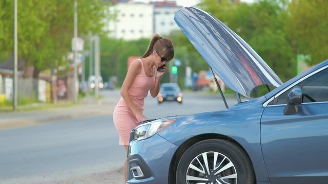 Helpless woman standing near her car with open bonnet calling road service for help. Young female driver having trouble with vehicle.