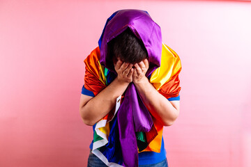 Young adult man wrapped in lgbtq flag covering his sad face. homophobia concept