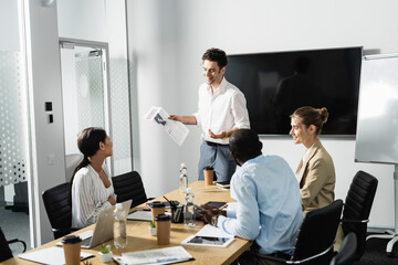 smiling businessman holding newspaper during meeting with multiethnic coworkers