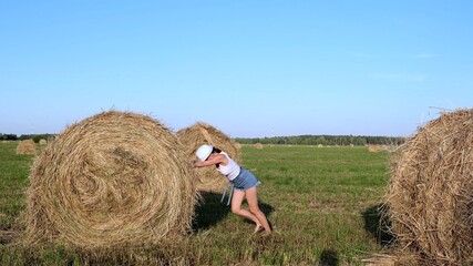 Naklejka na ściany i meble beautiful young girl in a hat pushes a haystack. Rural area. Feminist strong woman concept