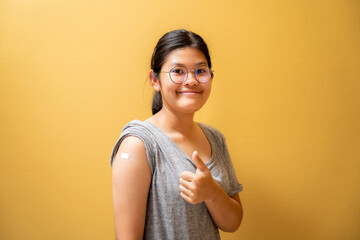 Portrait of asian teen girl giving thumbs up after getting covid-19 vaccine injection and showing bandage on his arm, Vaccinated - obrazy, fototapety, plakaty