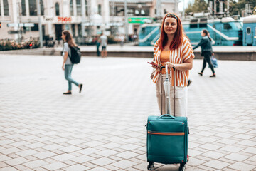 Young caucasian girl in stylish clothes with a smartphone and carry-on travel luggage stands at the...