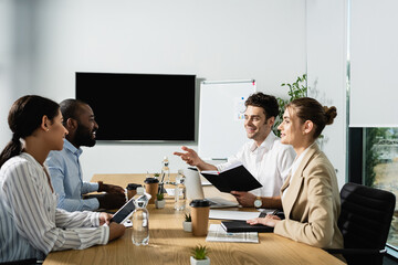 joyful businessman with notebook pointing with hand while talking to multiethnic colleagues