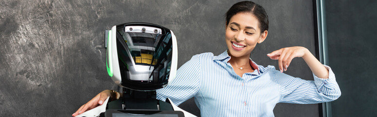 cheerful african american businesswoman pointing at robot in office, banner
