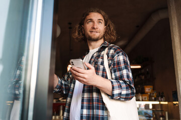 Young white cafe worker in plaid shirt using cellphone indoors
